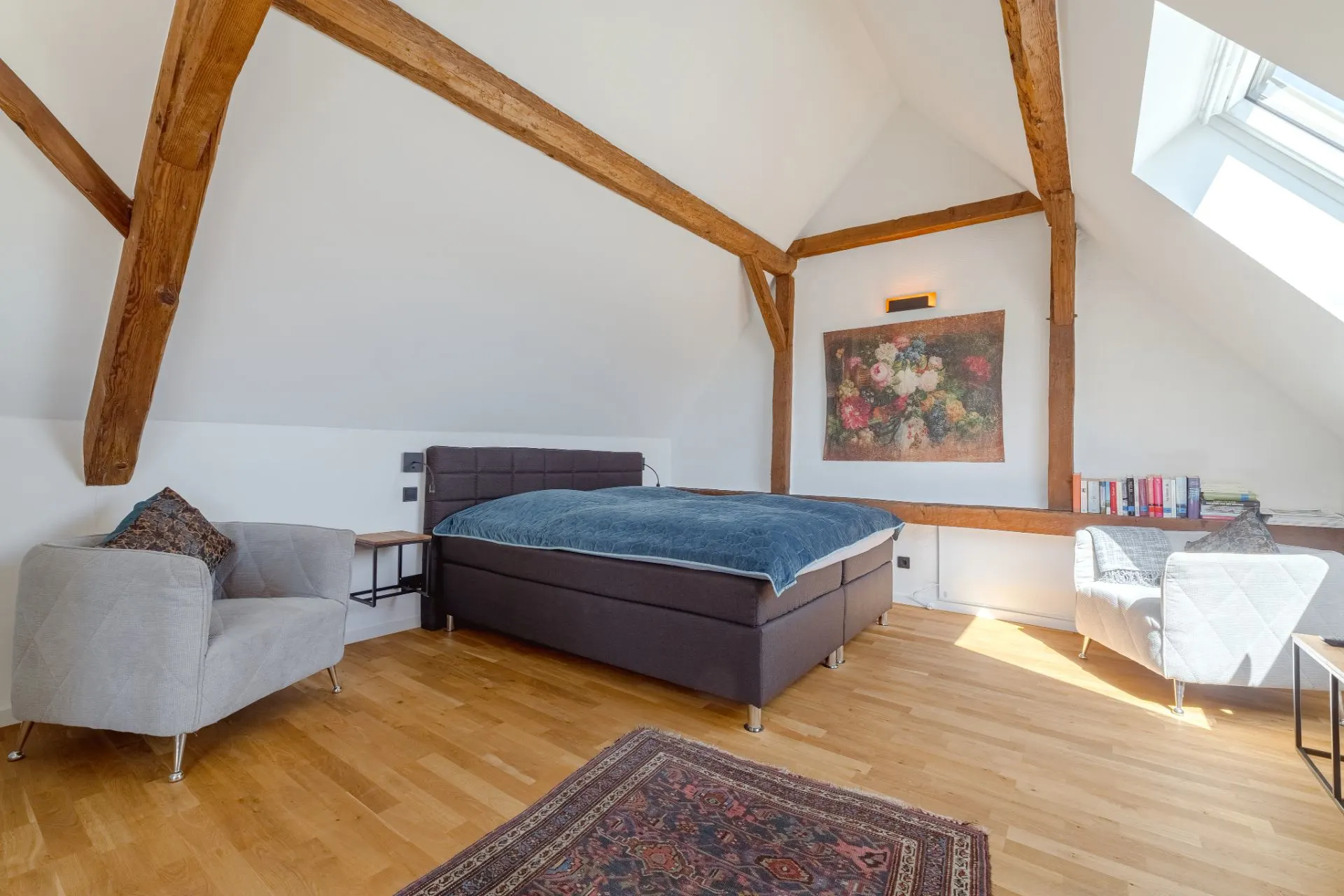 Helles Schlafzimmer mit Holzbalken, Doppelbett, zwei Sesseln und Teppich auf Holzfußboden.