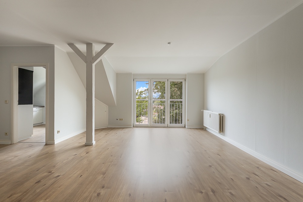 Helles, leeres Wohnzimmer mit Holzfußboden und großen Fenstertüren zum Balkon.