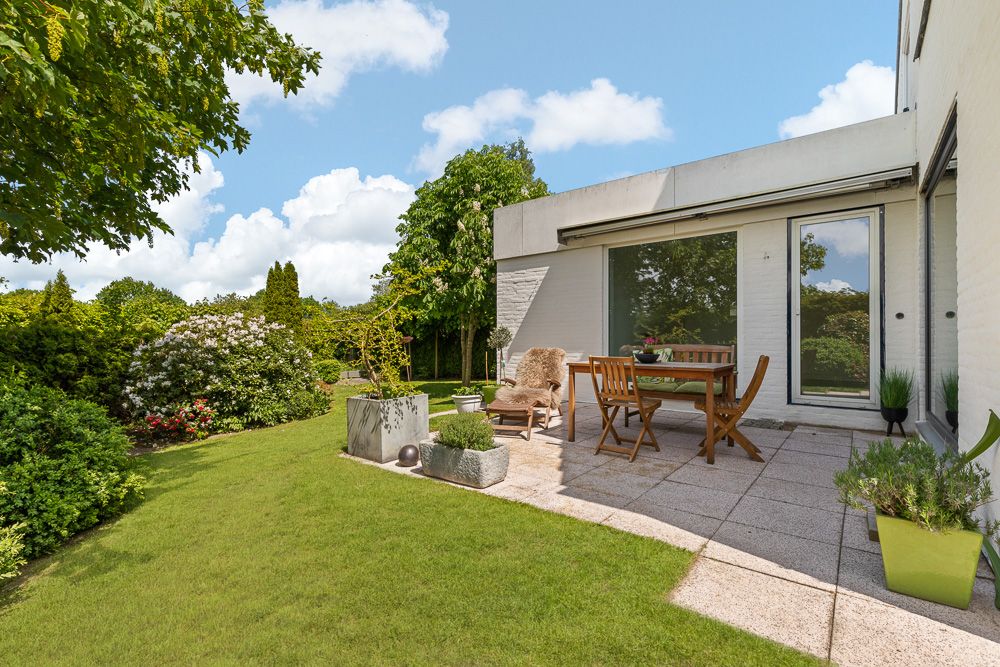 Moderne Terrasse mit Holzmöbeln, umgeben von grünem Garten und Bäumen bei sonnigem Wetter.