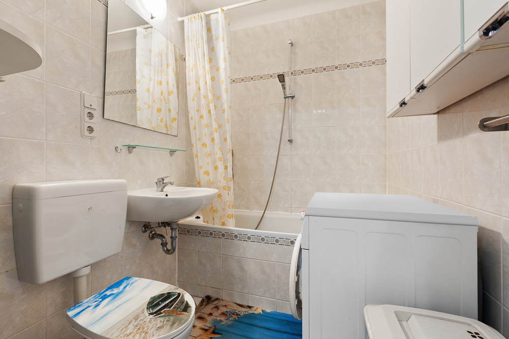 Kleines Bad mit Waschmaschine, Toilette, Waschbecken, Badewanne mit Duschvorhang und Wandspiegel.