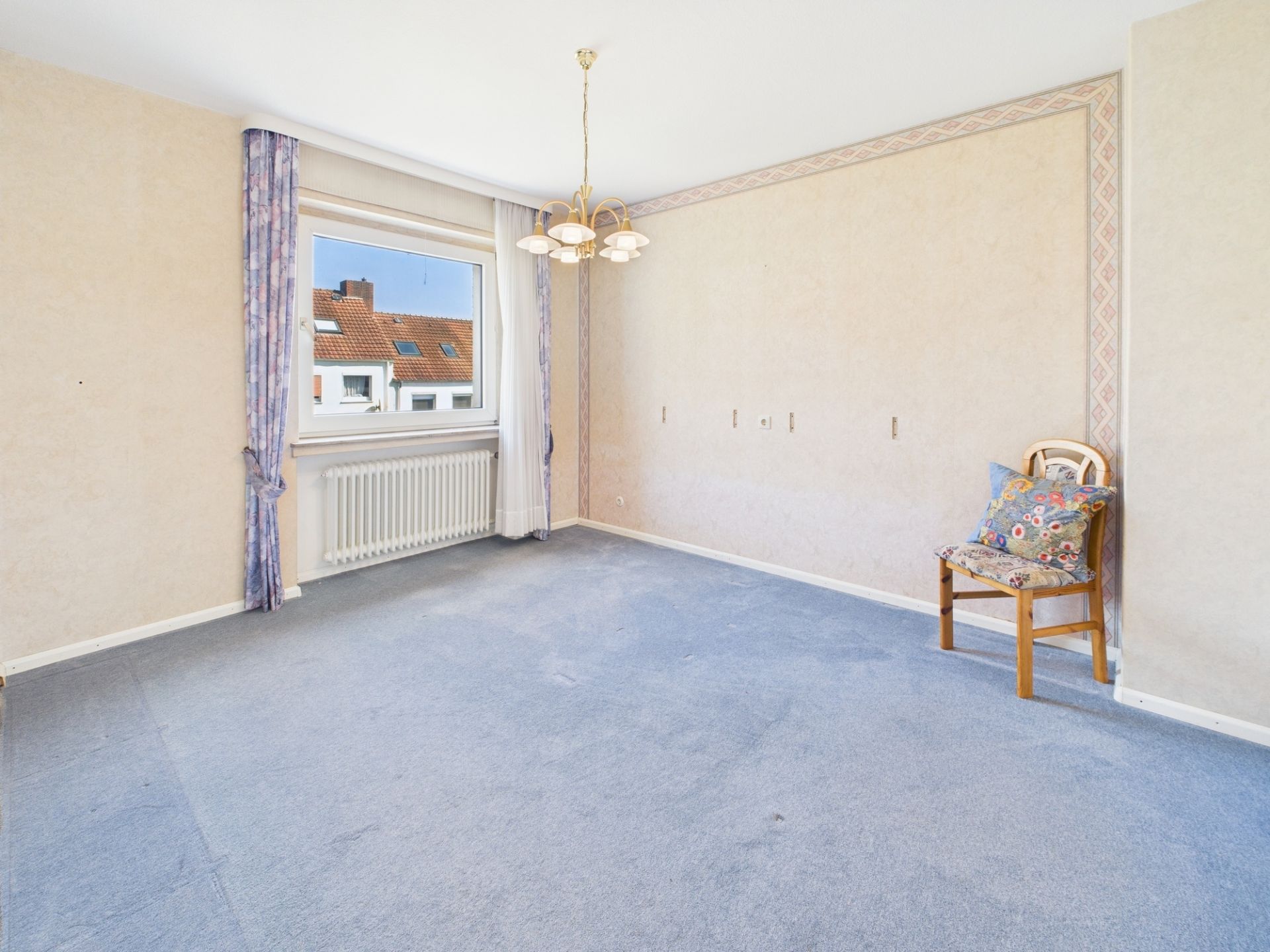 Helles Zimmer mit Teppichboden, Fenster, Vorhängen, Heizkörper und Stuhl mit Kissen.