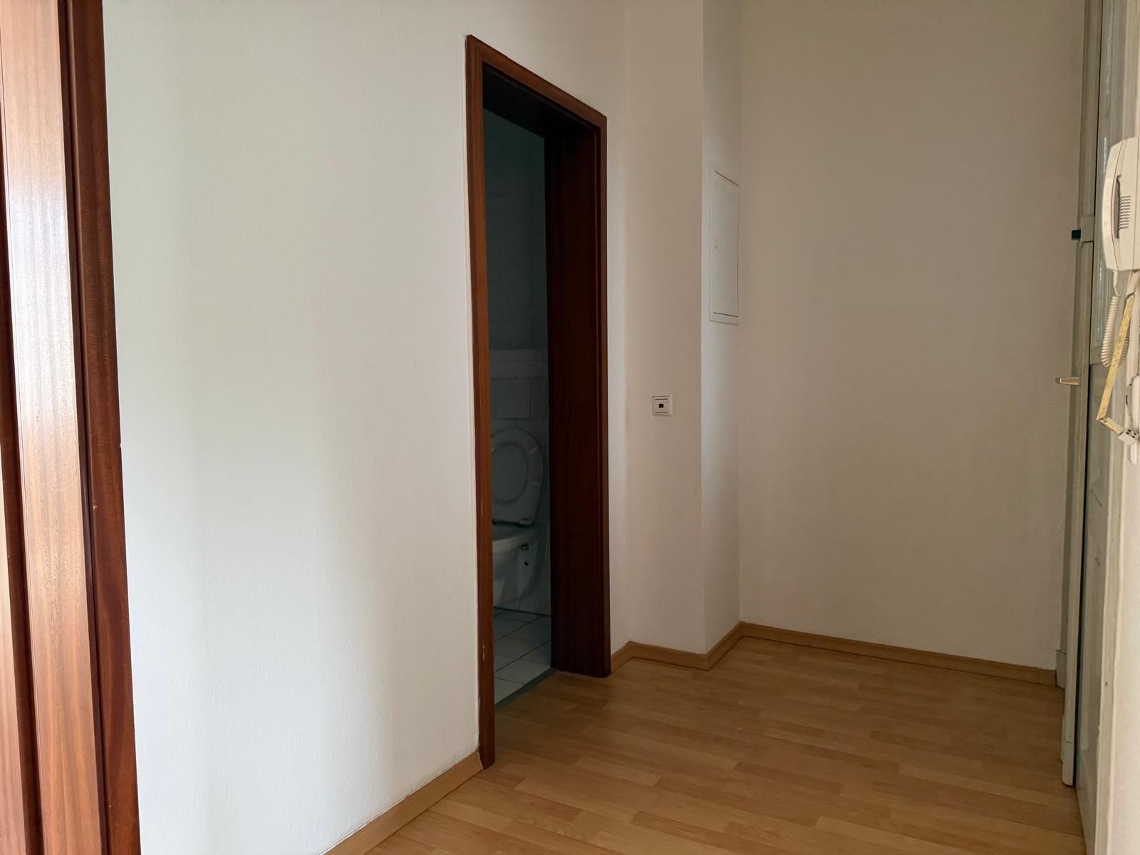 Flur mit Holzfußboden, weißer Wand und offener Tür zu einem Badezimmer mit Toilette.