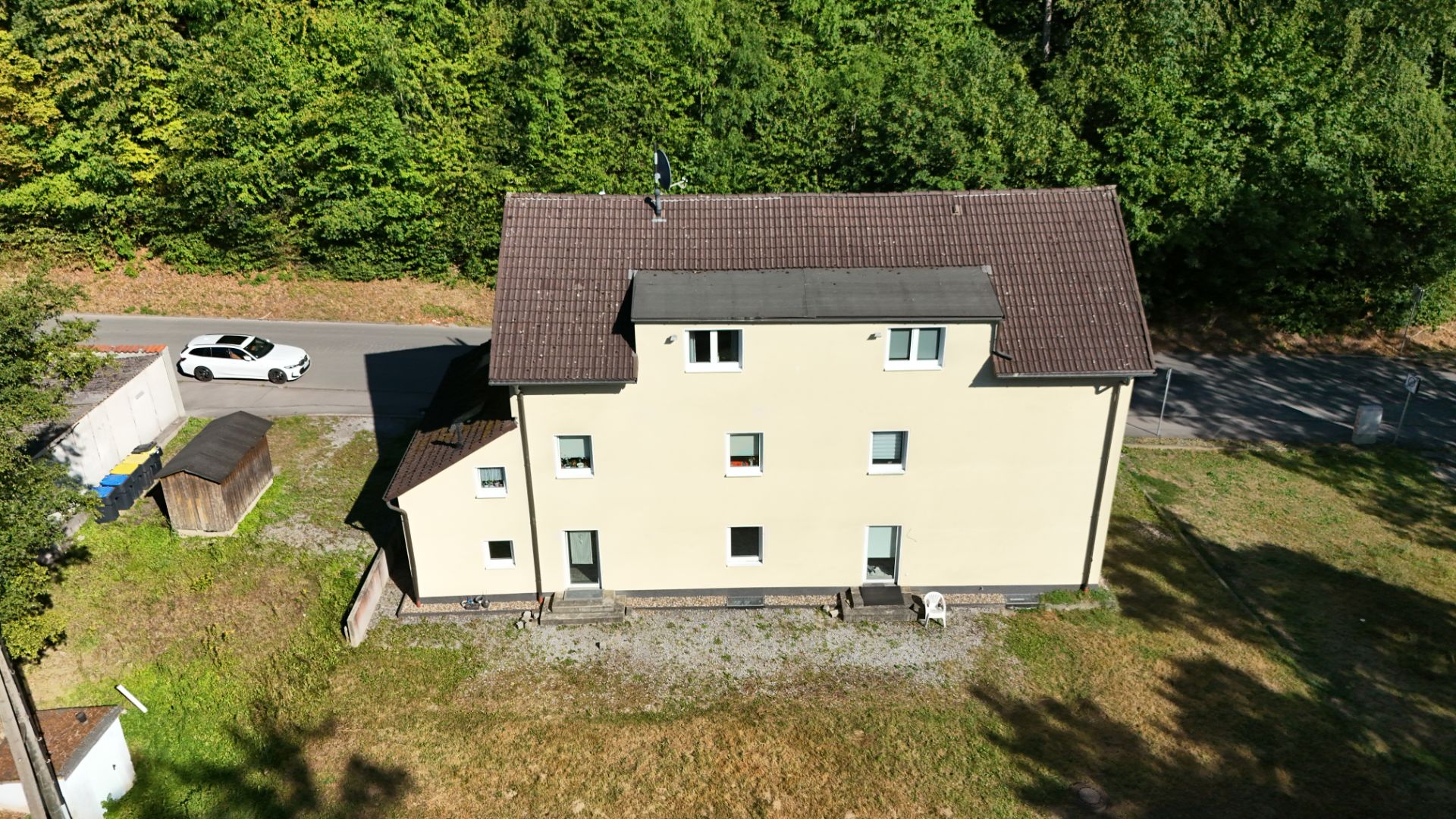 Beiges Wohnhaus mit Satteldach, umgeben von Bäumen und Rasen, Straße und Auto im Hintergrund.