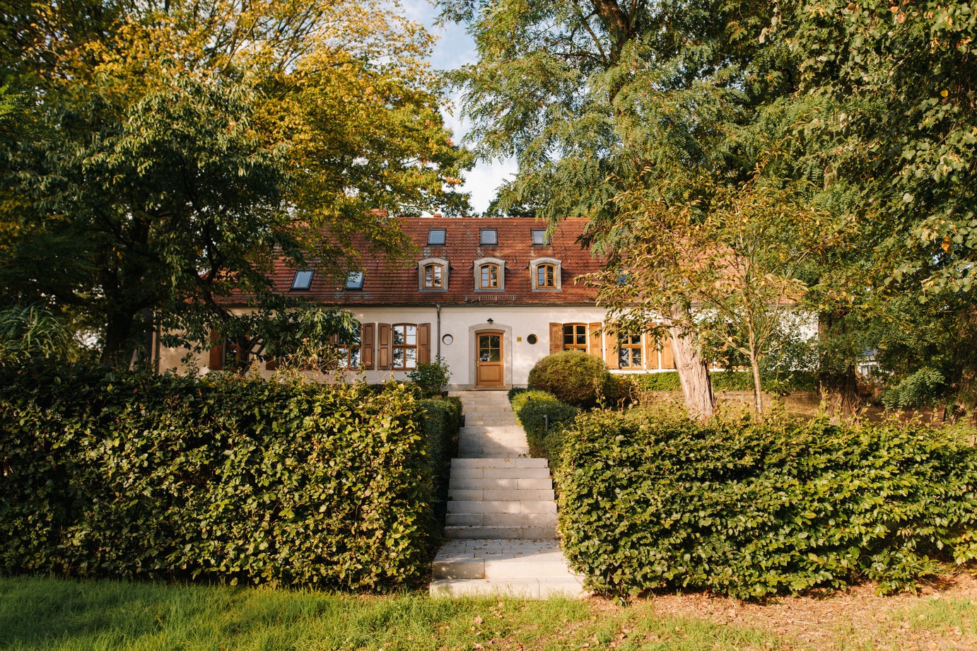 Wohnhaus mit rotem Dach, mehreren Fenstern und Treppe, umgeben von Bäumen und Hecken.