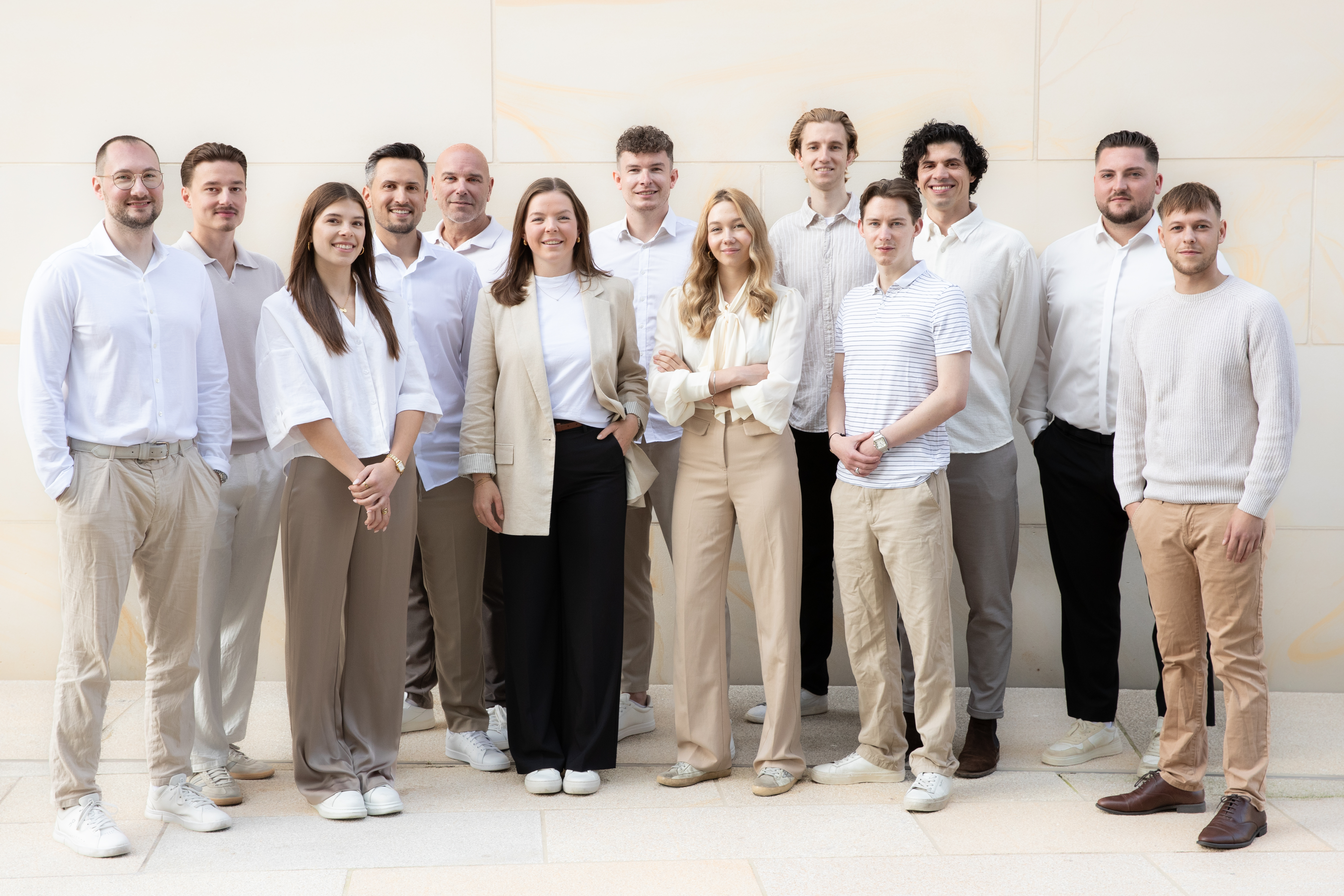 Gruppenfoto von dreizehn Menschen in heller Kleidung vor einer hellen Wand.
