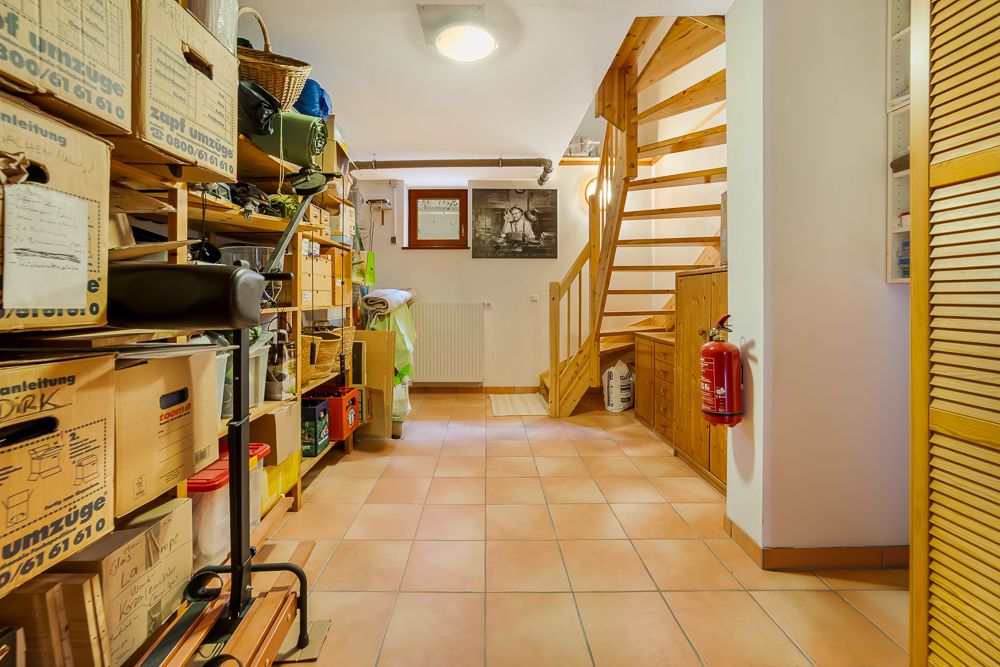 Kellerraum mit Regalen voller Kartons, Holztreppe und Feuerlöscher an der Wand.