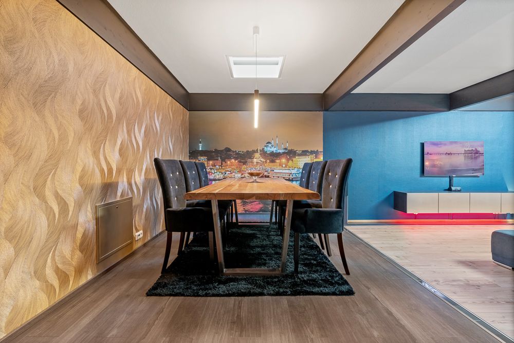 Essbereich mit Holztisch, sechs Stühlen und Wandbild, daneben Wohnzimmer mit Fernseher.
