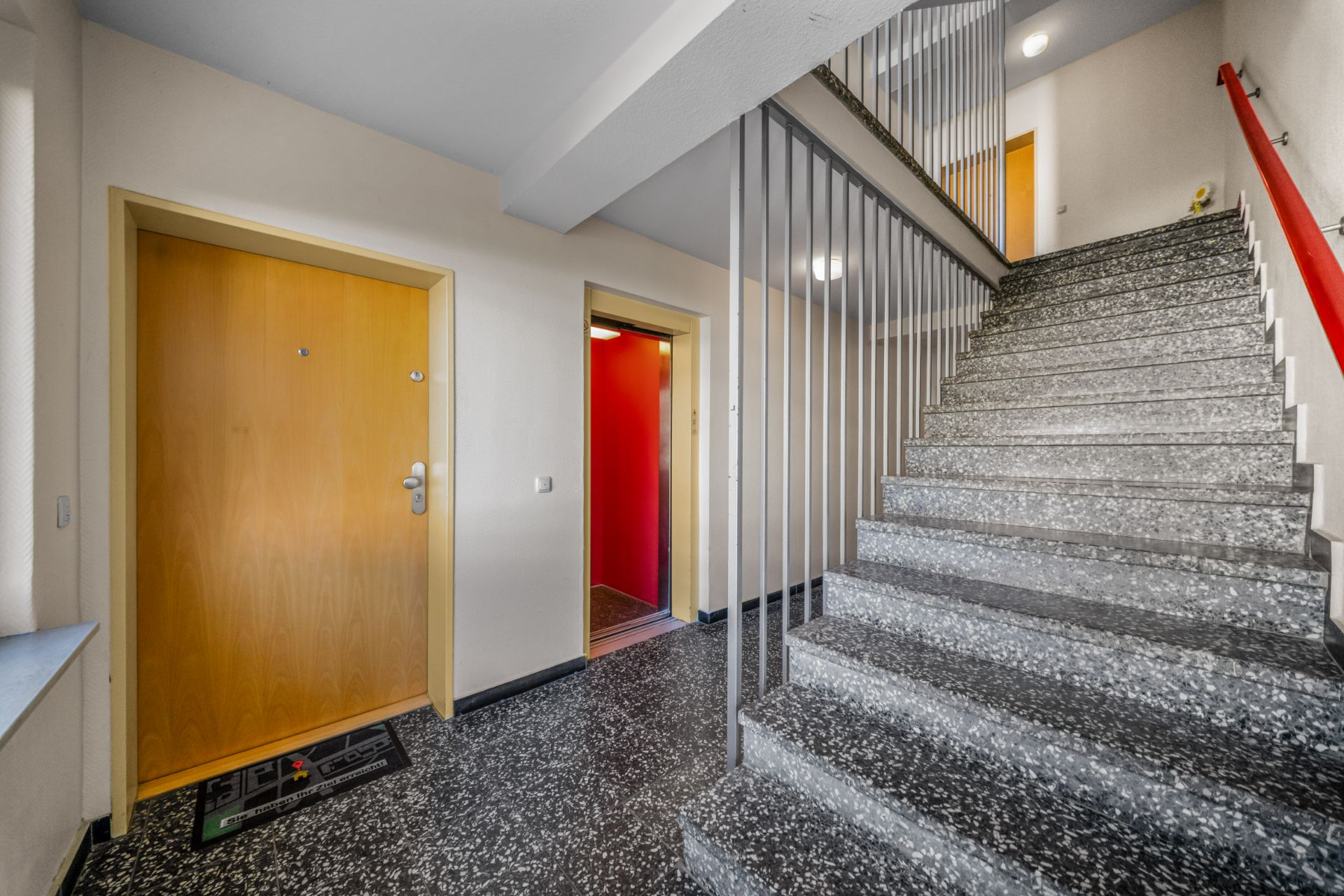 Treppenhaus mit grauer Treppe, Geländer und zwei Holztüren in einem Wohngebäude.