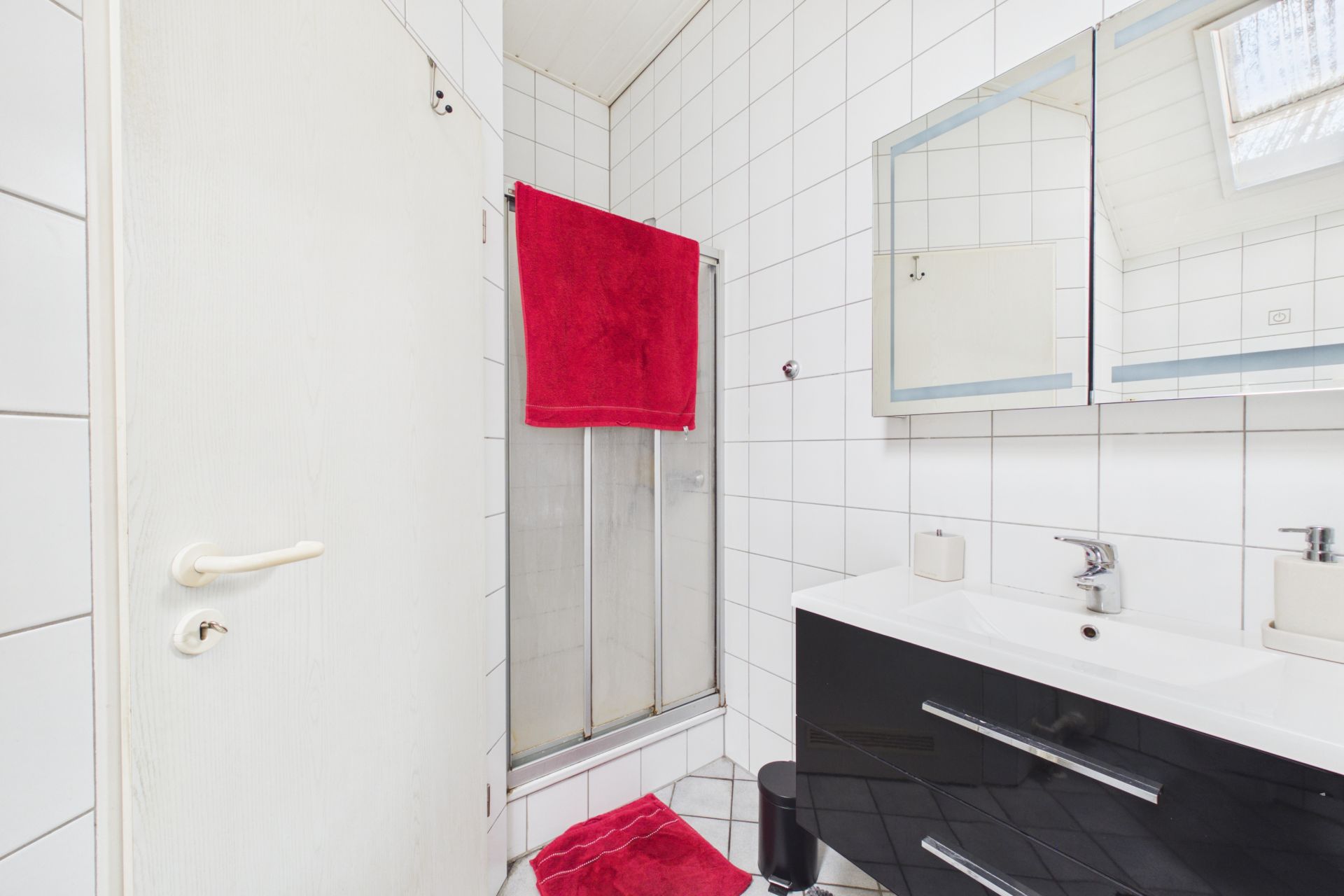Helles Badezimmer mit Dusche, rotem Handtuch, Waschbecken und Spiegel.