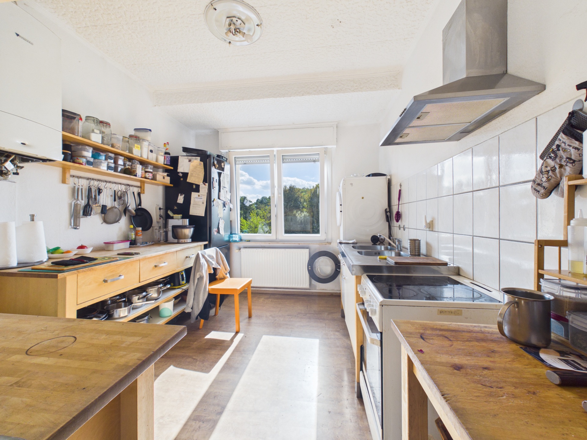 Helle Küche mit Holzmöbeln, großem Fenster, Herd, Kühlschrank und Regalen mit Küchenutensilien.