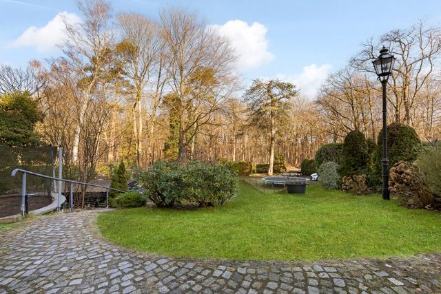 Gepflasterter Weg mit grüner Rasenfläche, Sträuchern und Laterne im Garten vor einem bewaldeten Hintergrund.