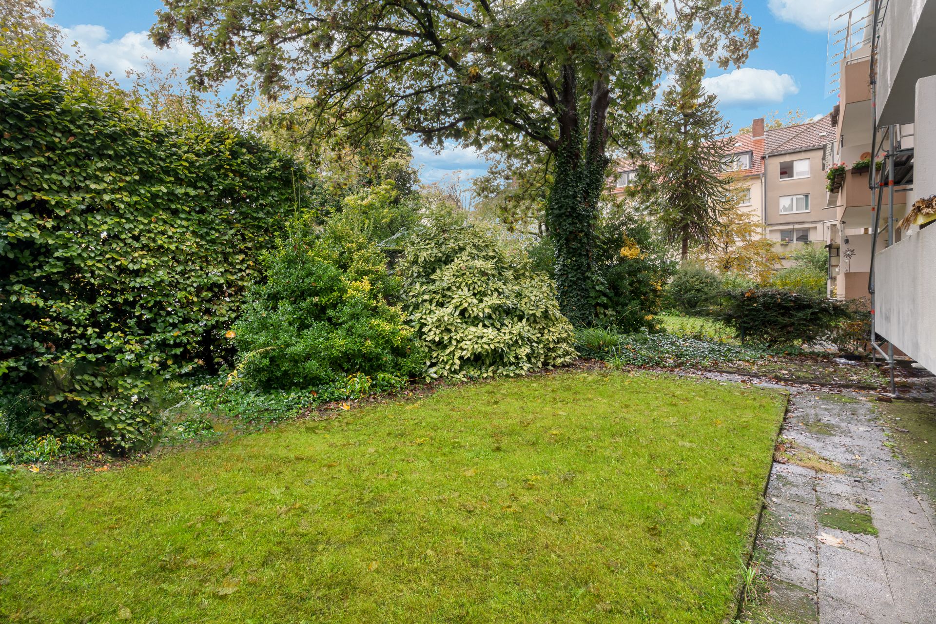 Grüner Garten mit Rasen, Sträuchern und Bäumen neben einem Wohngebäude.