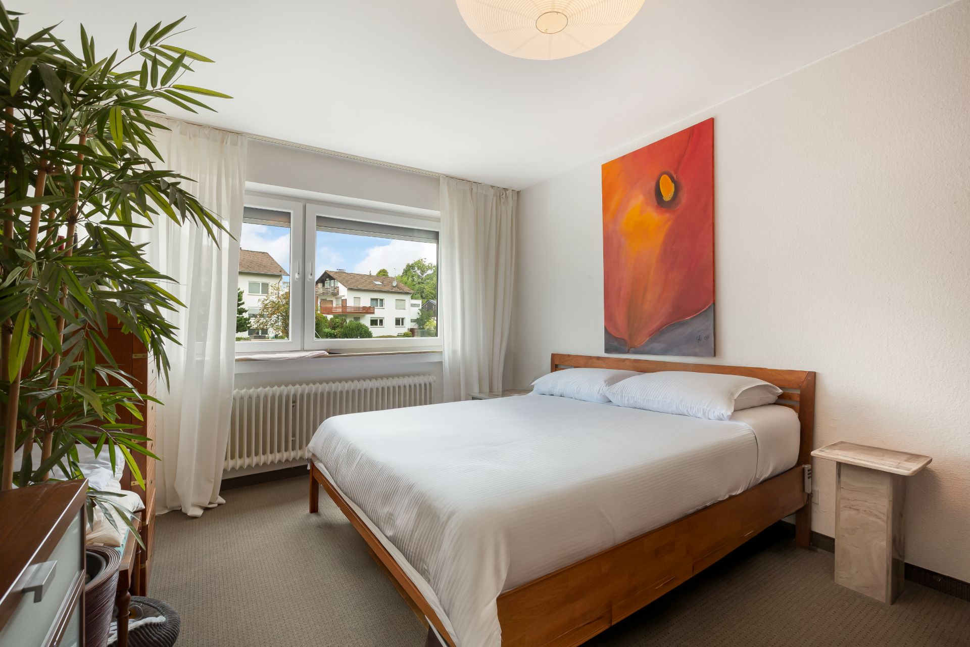 Schlafzimmer mit Doppelbett, großem Fenster, weißem Vorhang und bunter Wandkunst.