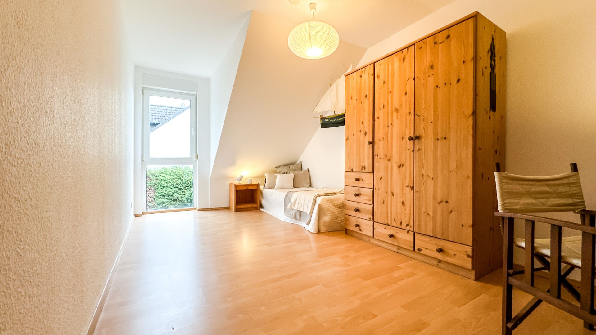 Helles Zimmer mit Holzboden, Einzelbett, großem Kleiderschrank, Stuhl und Fenster.