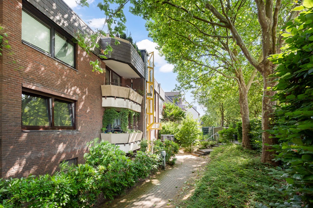 Außenansicht eines Wohngebäudes mit Balkonen und grünem Gartenweg unter Bäumen.