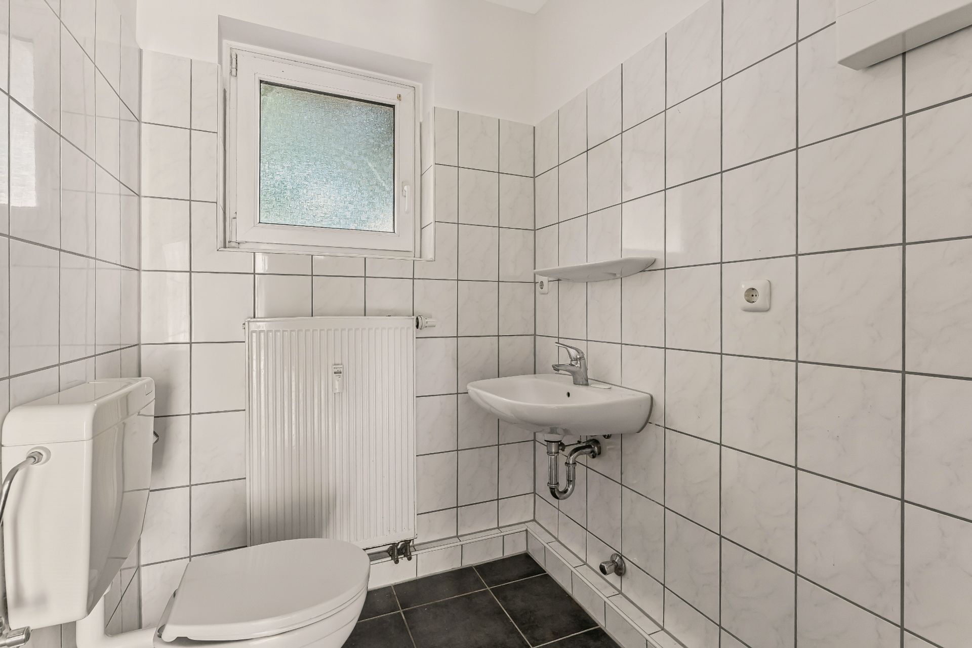 Kleines Badezimmer mit WC, Waschbecken, Heizung und Fenster, weiße Fliesen an den Wänden.