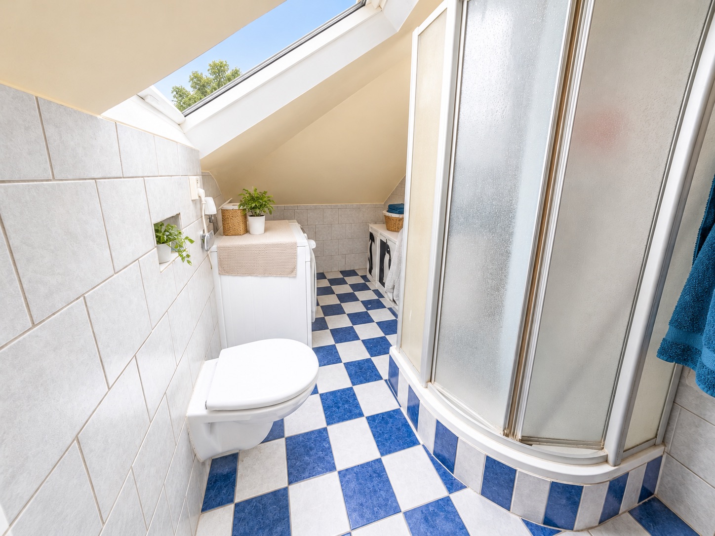 Kleines Badezimmer mit Schrägdach, Dusche, Toilette und blau-weiß karierten Fliesen.