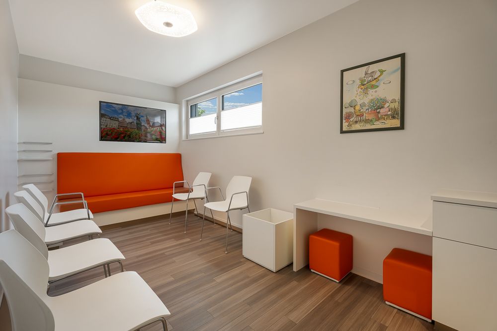 Heller Raum mit weißen Stühlen, orangefarbener Sitzbank, Fenstern und Wandbildern.