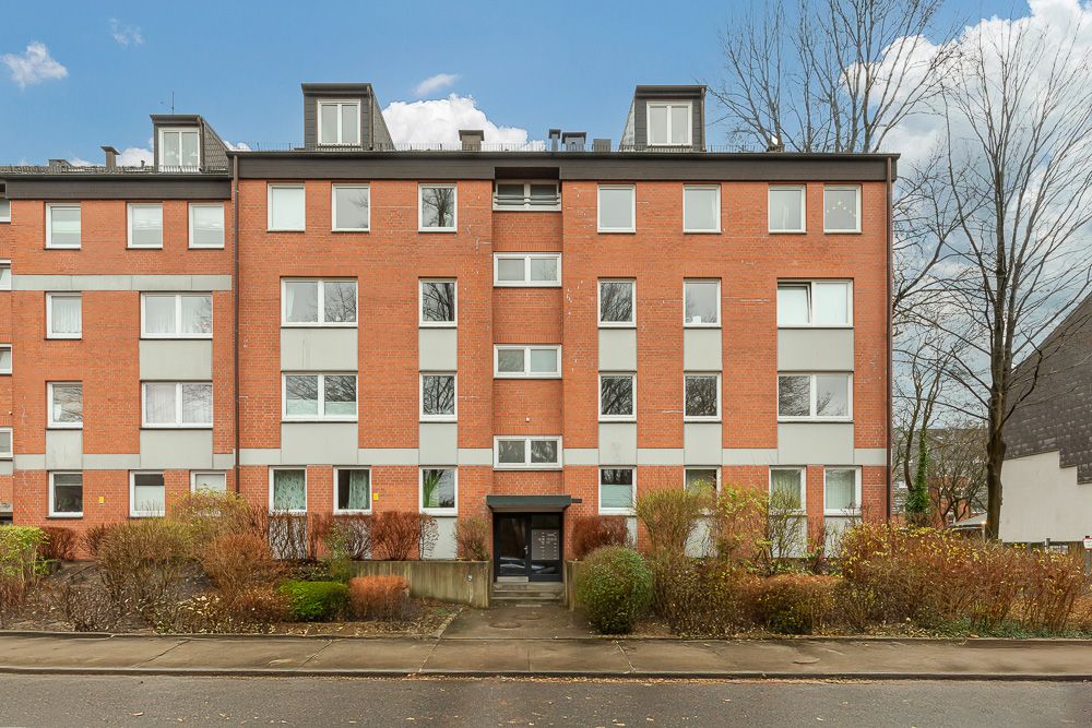 Mehrstöckiges Wohnhaus mit roter Klinkerfassade und vielen Fenstern, vorne Büsche und Gehweg.