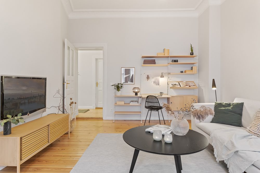 Helles Wohnzimmer mit Sofa, rundem Tisch, TV, Regalwand und Schreibtisch auf Holzboden.