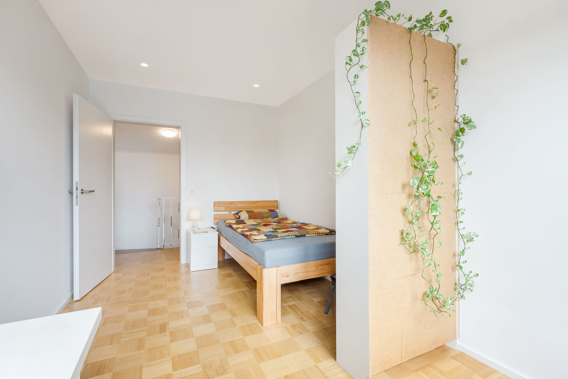 Helles Schlafzimmer mit Holzbett, Nachttisch und hölzernem Boden. An der Wand hängt eine grüne Ranke.