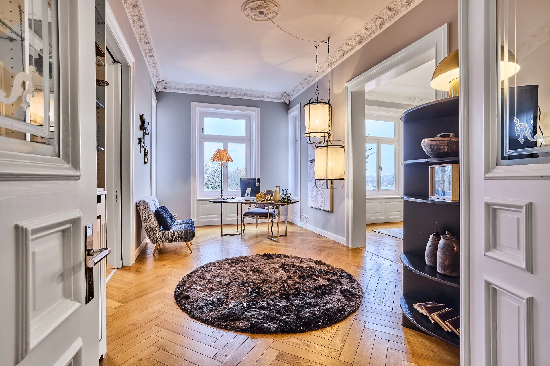 Heller Wohnraum mit rundem Teppich, Schreibtisch am Fenster und Regal rechts.