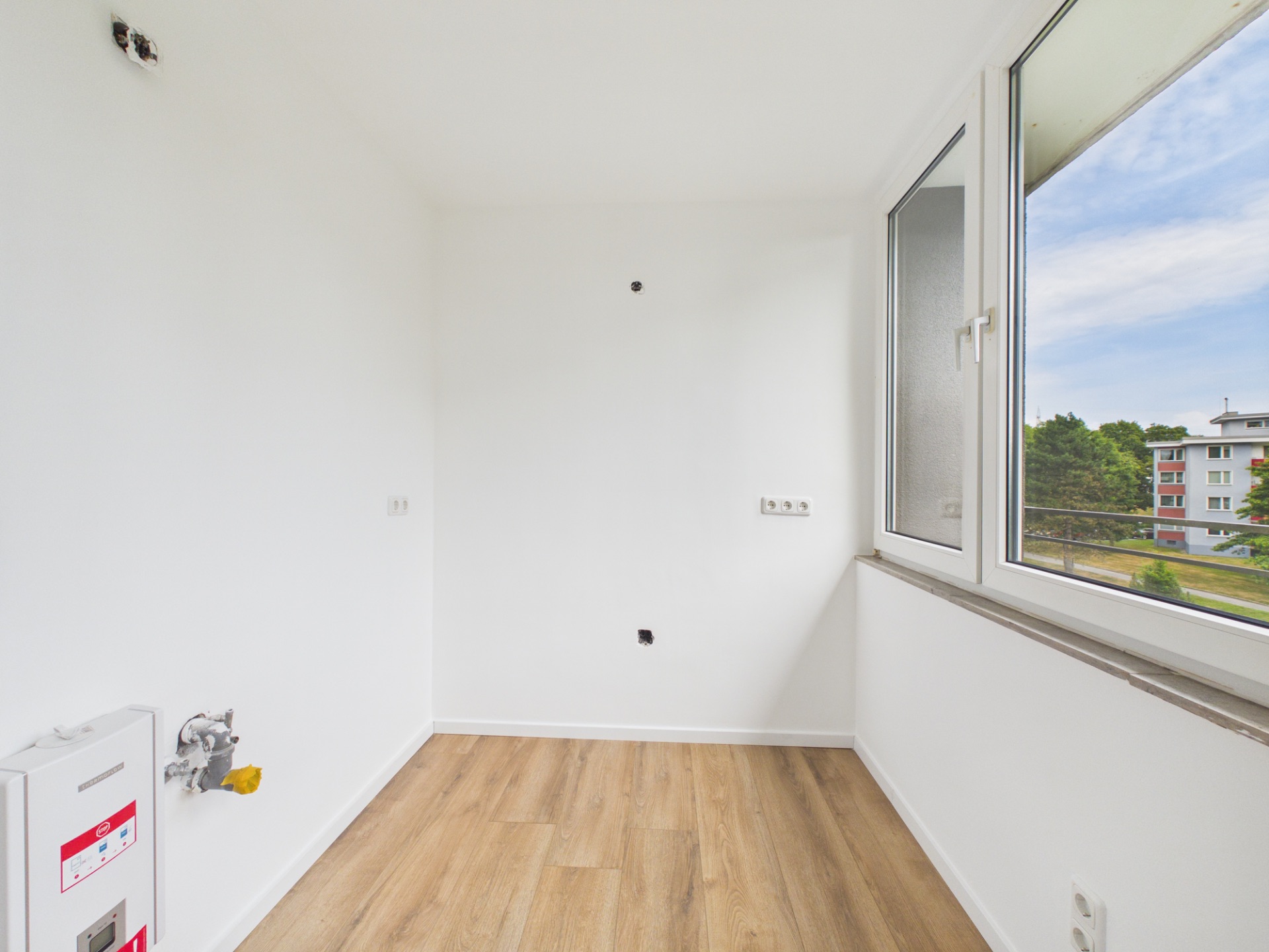 Leerer Raum mit weißer Wand, Fenster und hellem Holzfußboden. Anschlüsse befinden sich an der Wand.