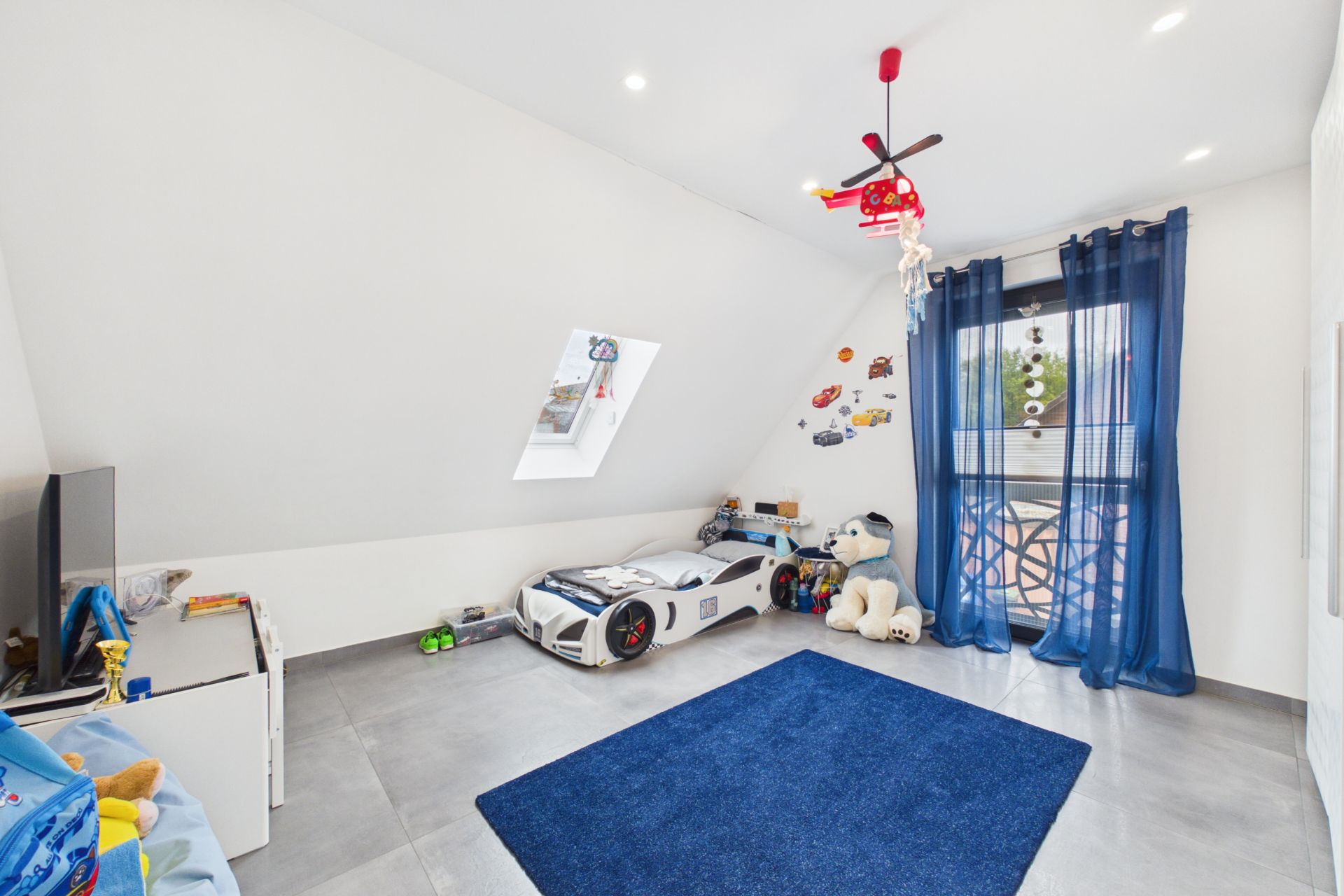 Kinderzimmer mit Auto-Bett, Plüschtieren, TV, blauem Teppich und Dachfenster.