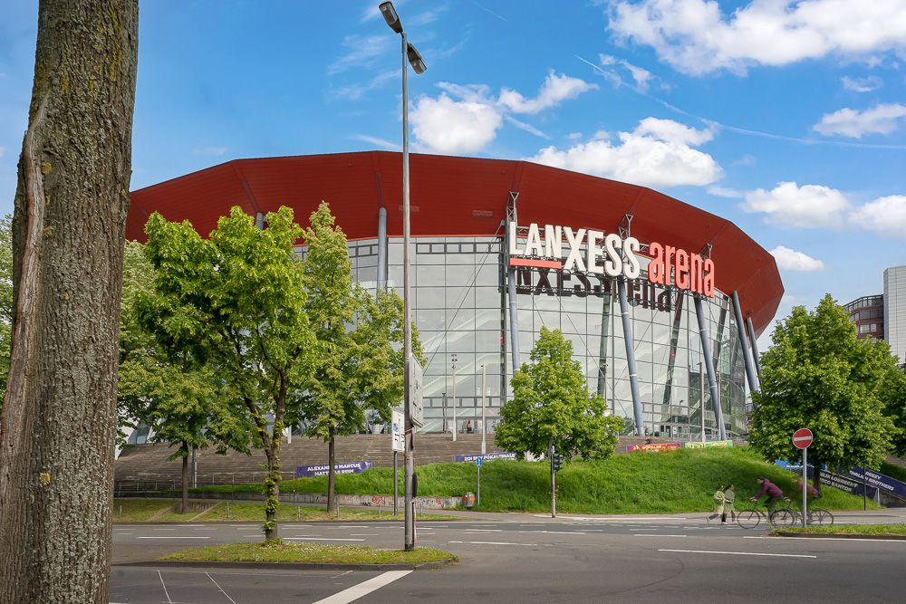 Große, moderne Arena mit Glasfassade und rotem Dach, Bäume und Straße im Vordergrund.
