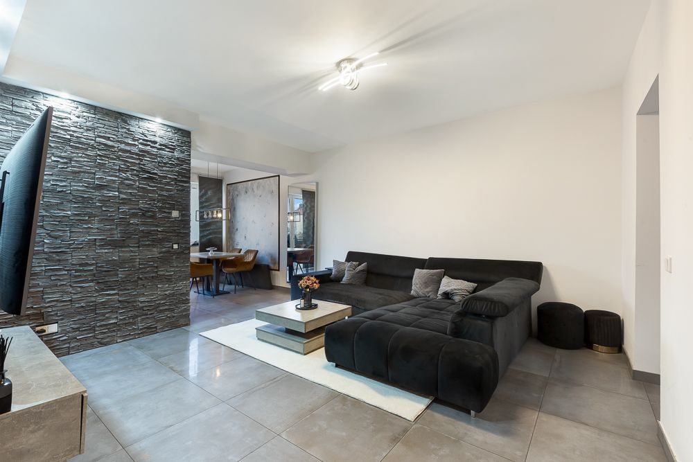 Modernes Wohnzimmer mit schwarzem Ecksofa, Couchtisch und Steinwand, hinten Essbereich sichtbar.