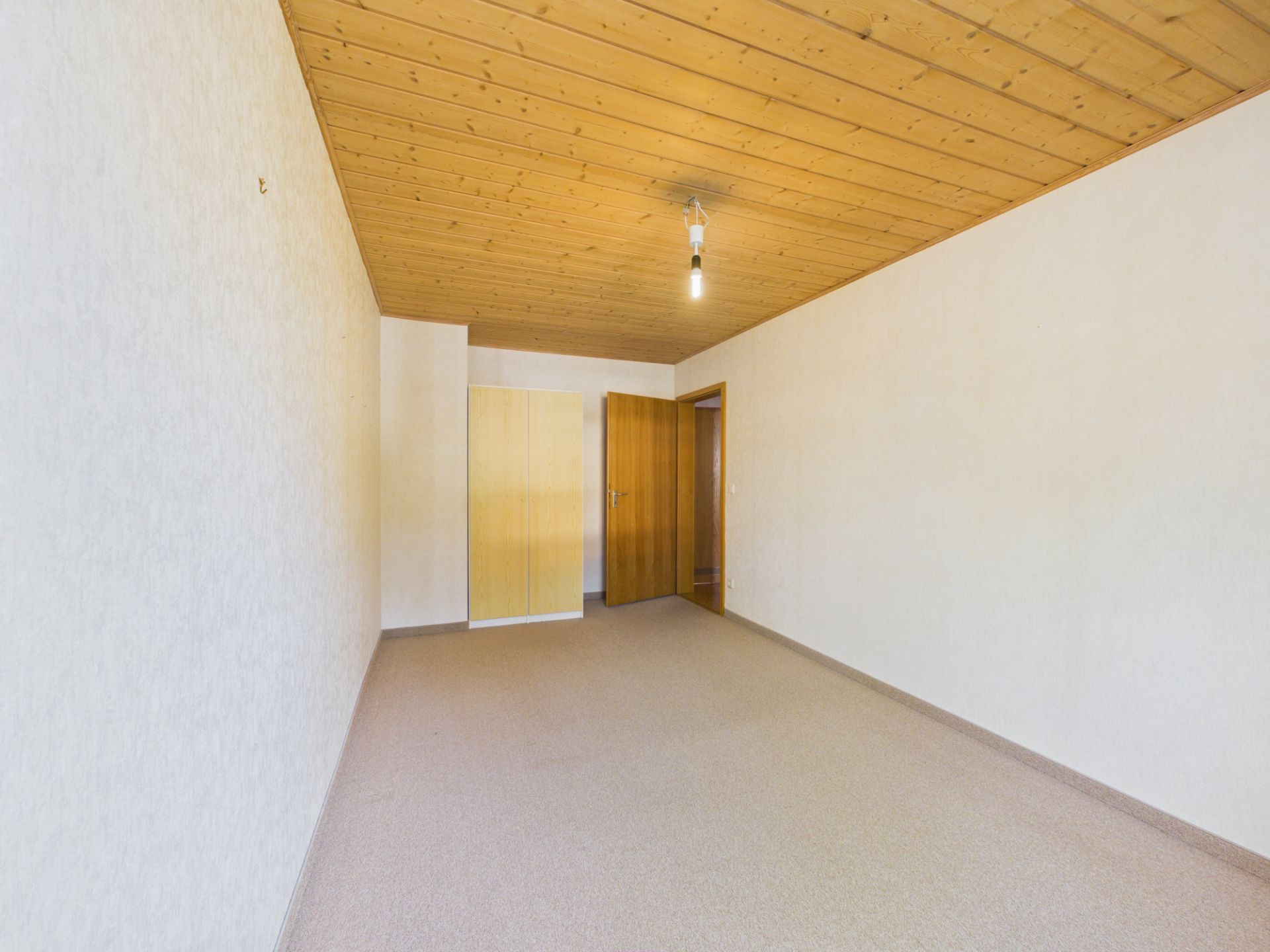 Leerer Raum mit Teppichboden, Holztür, Holzdecke und einer einfachen Lampe.
