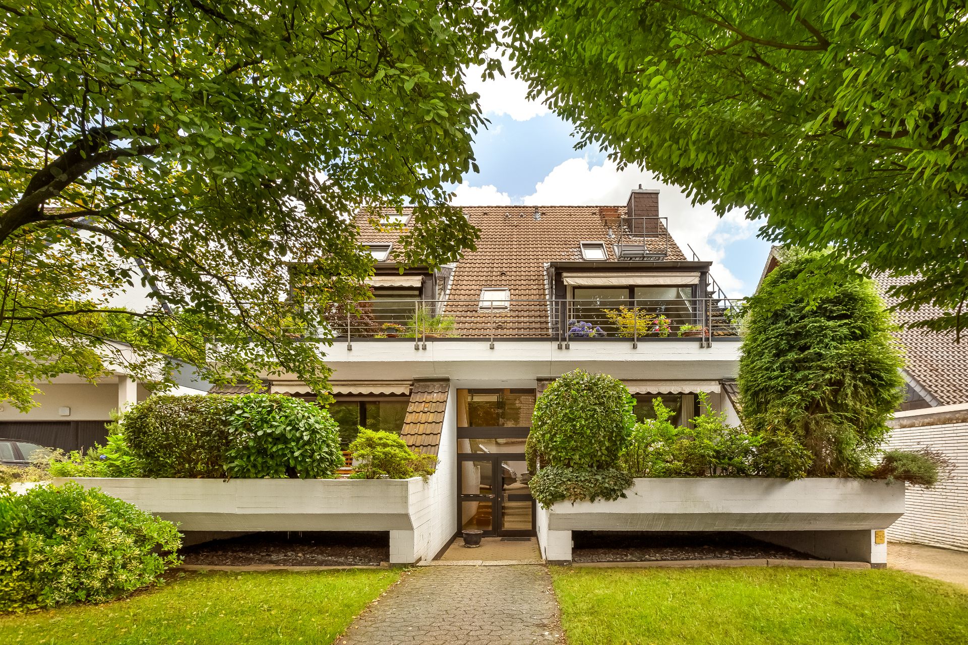 Mehrfamilienhaus mit Balkon, bepflanztem Vorgarten und Gehweg, umgeben von Bäumen.