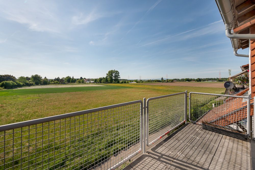 Balkon mit Metallgeländer und Blick auf weite grüne Felder unter blauem Himmel.