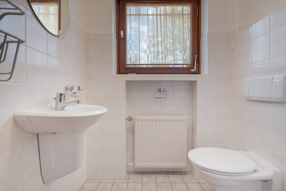 Kleines Bad mit Waschbecken, WC, Fenster und Heizkörper, weiße Wandfliesen.