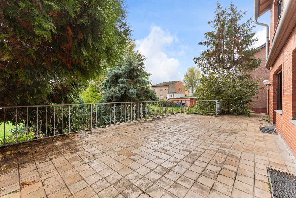 Große geflieste Terrasse mit Metallgeländer, daneben Bäume und Sträucher.