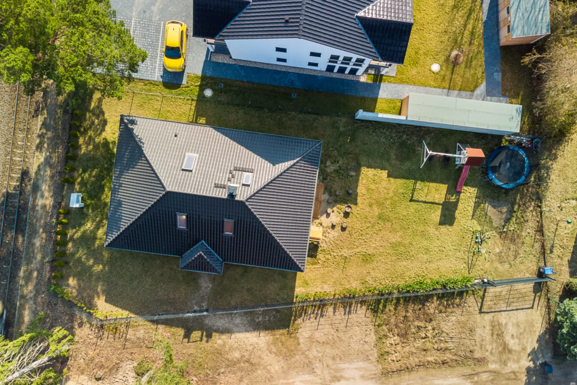 Luftaufnahme eines Hauses mit Garten, Spielplatz und Trampolin neben einer Einfahrt mit gelbem Auto.