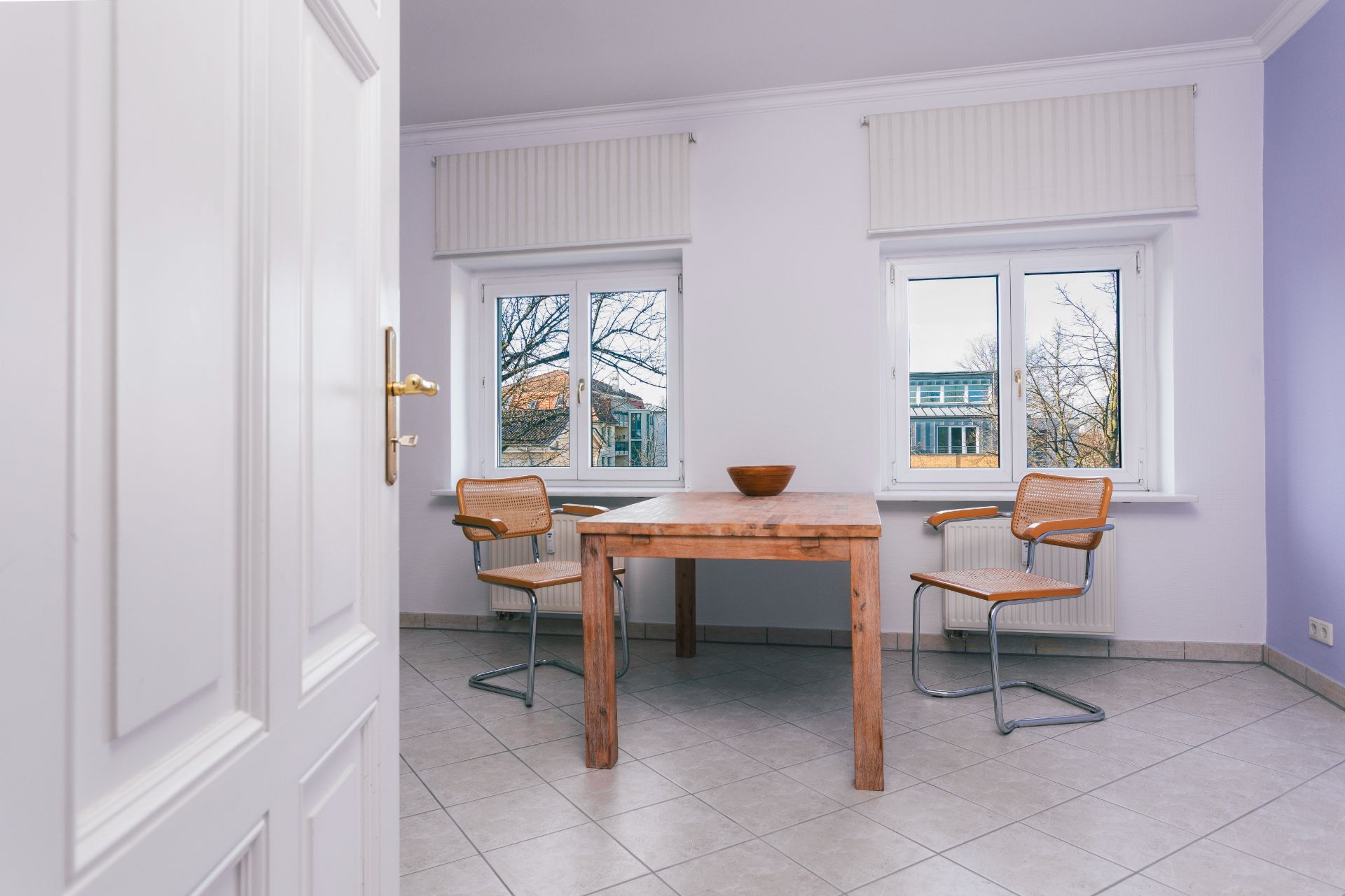 Helles Zimmer mit Holztisch, zwei Stühlen und zwei Fenstern mit Blick nach draußen.