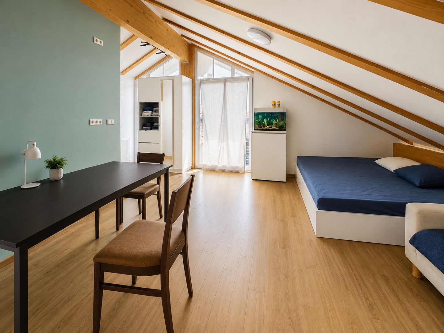 Helles Dachzimmer mit Bett, Tisch, Stuhl, Schrank und Aquarium auf Holzfußboden.