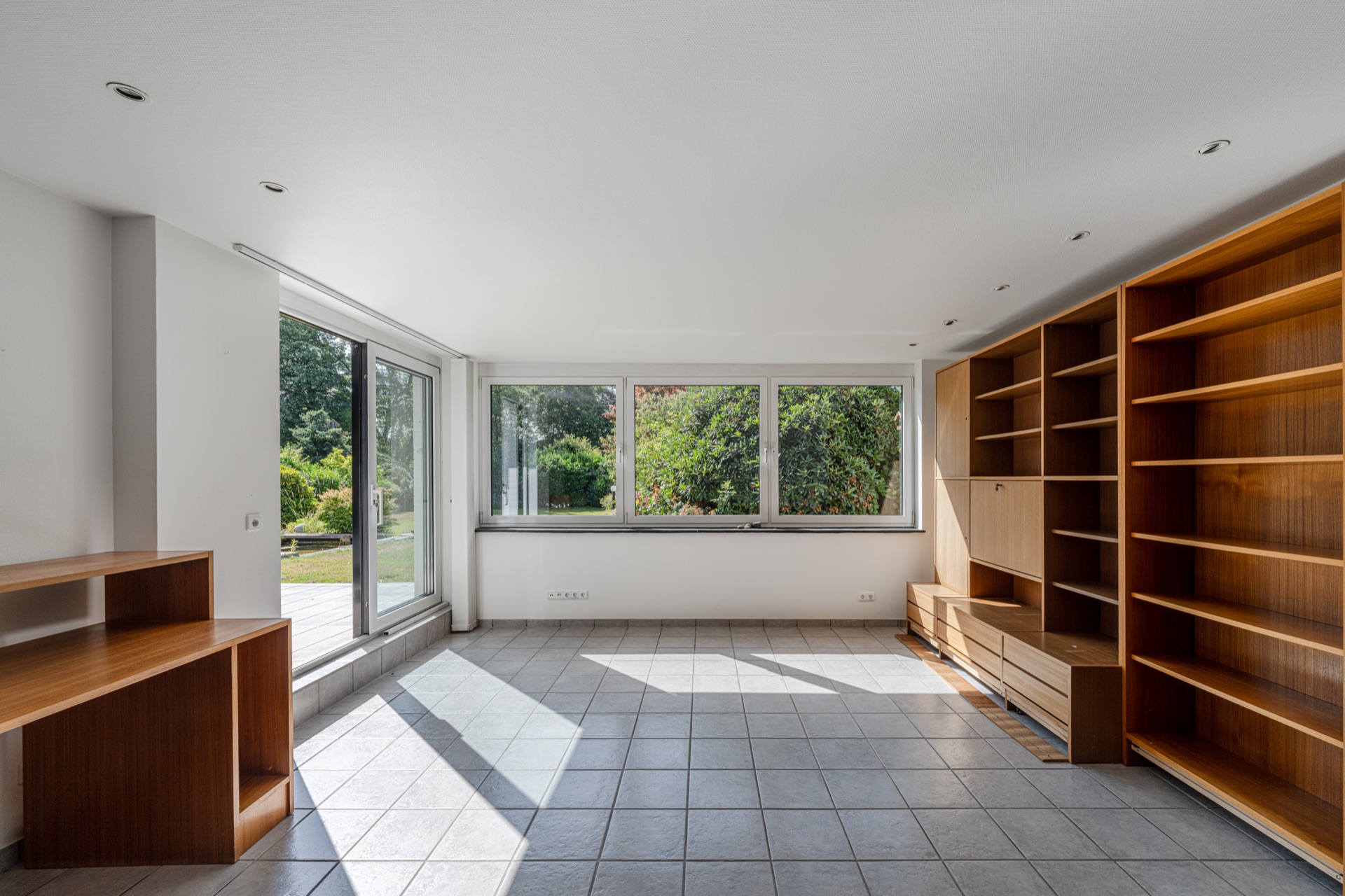 Heller Raum mit großen Fenstern, Fliesenboden und Holzregalen an der rechten Wand.