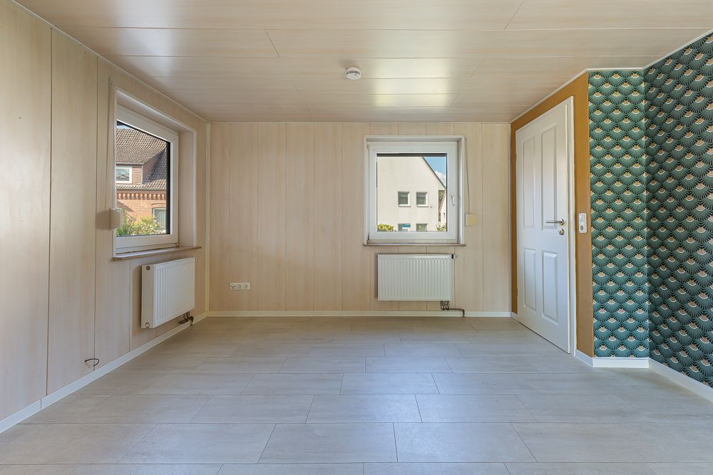 Leerer Raum mit zwei Fenstern, Heizkörpern, einer Tür und gemusterten Tapeten an einer Wand.