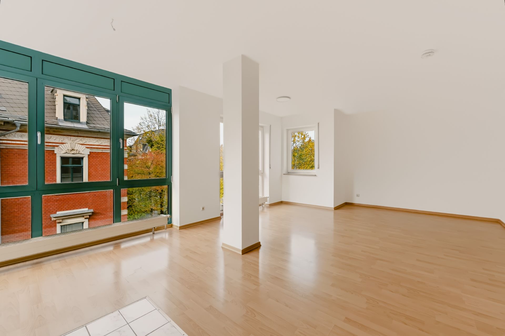 Helles, leeres Zimmer mit großen Fenstern, Holzfußboden und weiß gestrichenen Wänden.