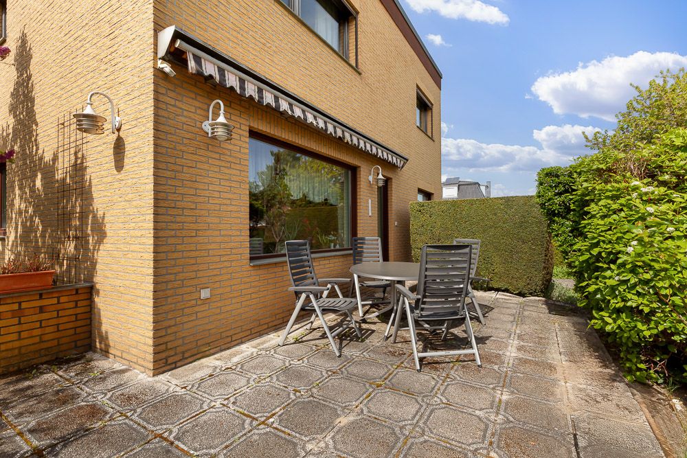 Gepflasterte Terrasse mit Tisch, vier Stühlen und Markise an einem Wohnhaus, umgeben von Hecken.