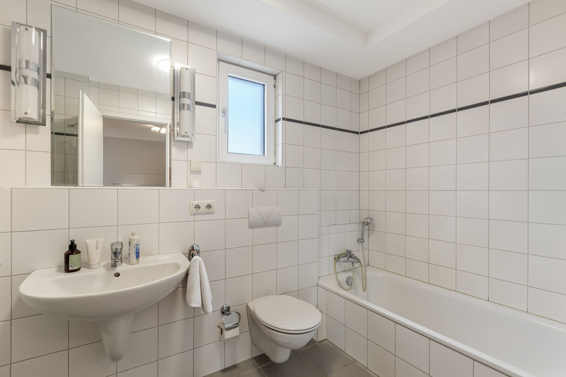 Modernes Badezimmer mit weißen Fliesen, Waschbecken, Spiegel, Toilette und Badewanne.
