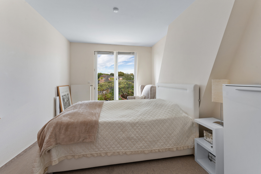 Helles Schlafzimmer mit Doppelbett, weißer Bettwäsche und Blick aus großen Fenstern auf einen Garten.