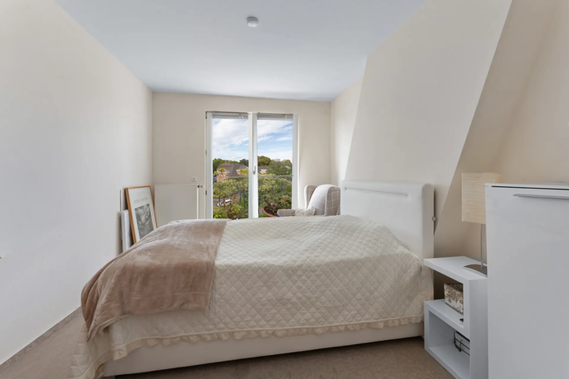 Helles Schlafzimmer mit Doppelbett, weißer Bettwäsche und Blick aus großen Fenstern auf einen Garten.