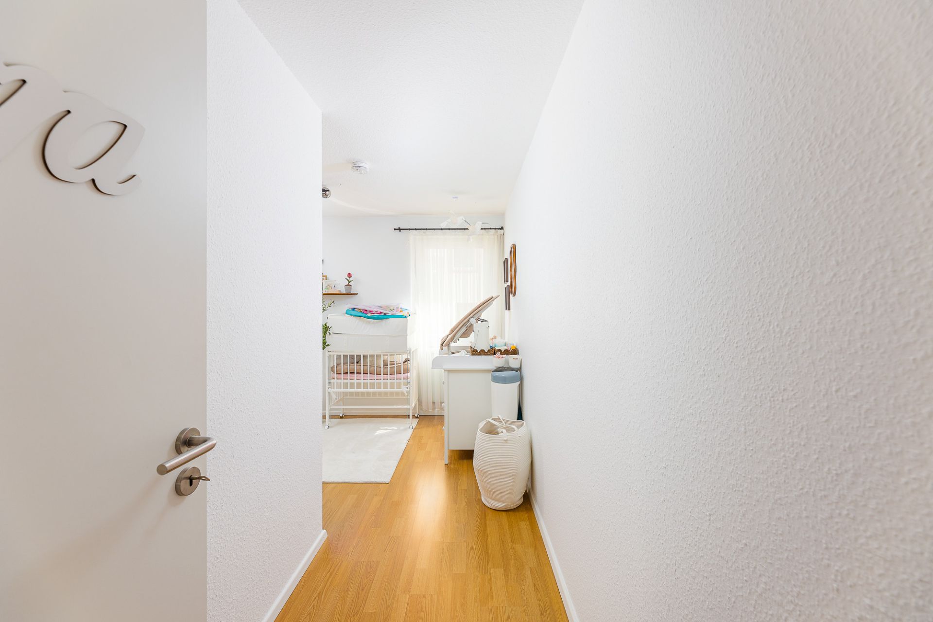 Heller Flur mit Blick in ein Kinderzimmer mit Gitterbett, Wickeltisch und Holzfußboden.