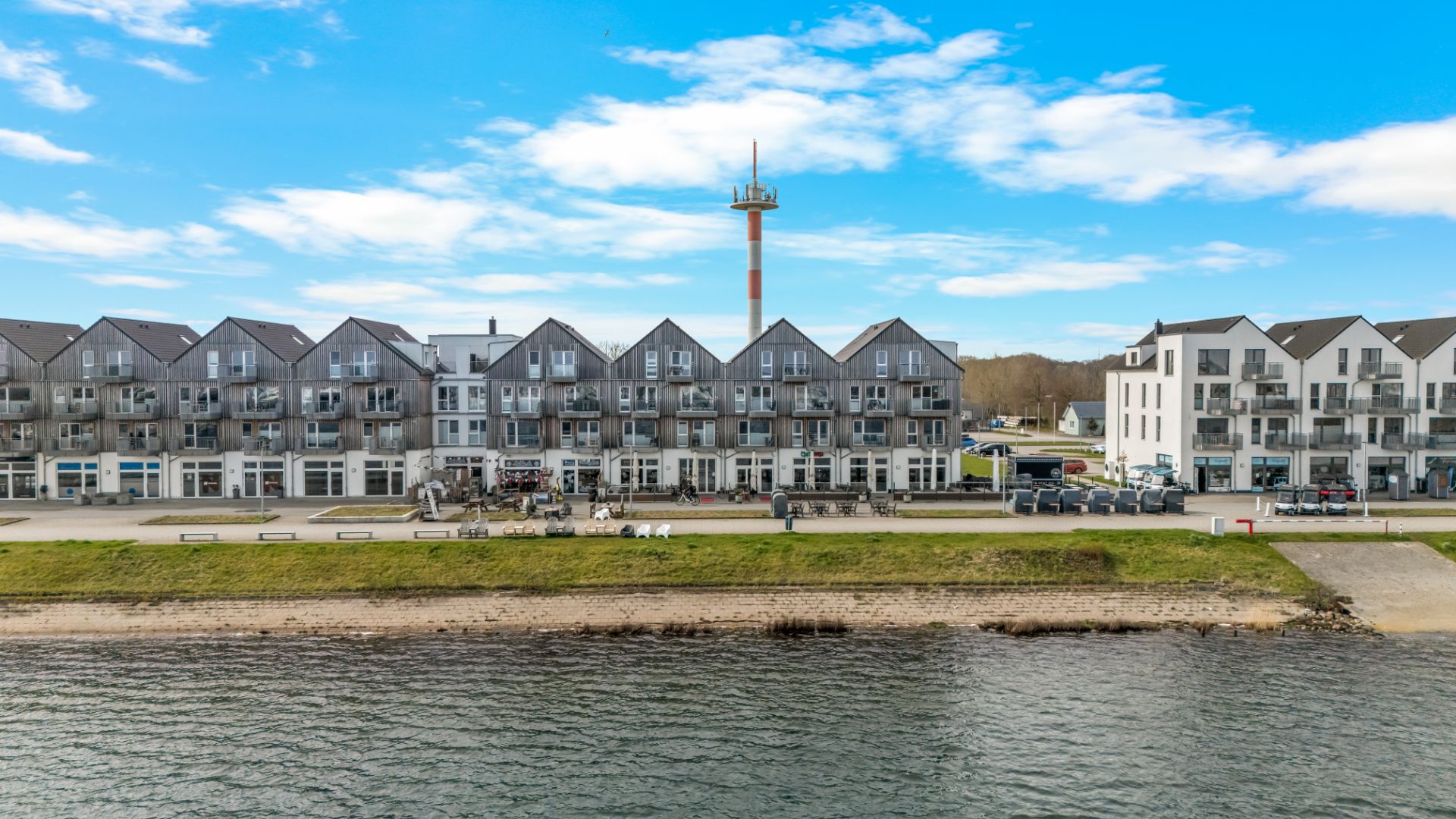 Mehrere moderne Wohnhäuser stehen an einer Uferpromenade mit Wasser im Vordergrund.