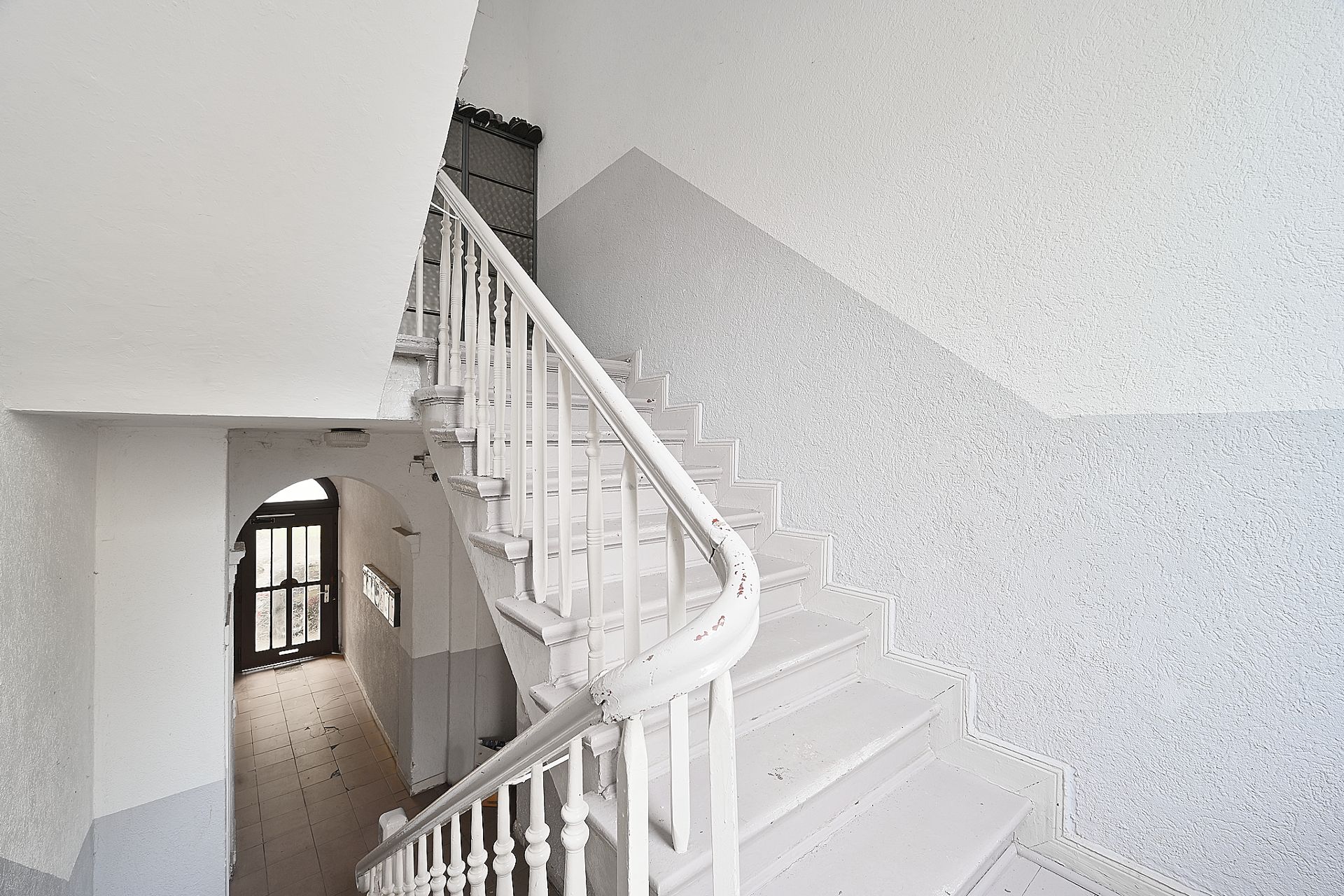 Helles Treppenhaus mit weißer Holztreppe und Handlauf, daneben ein Flur mit Eingangstür.