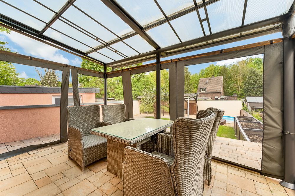 Überdachte Terrasse mit Glasdach, Rattanmöbeln und Glastisch, Blick in einen grünen Garten.