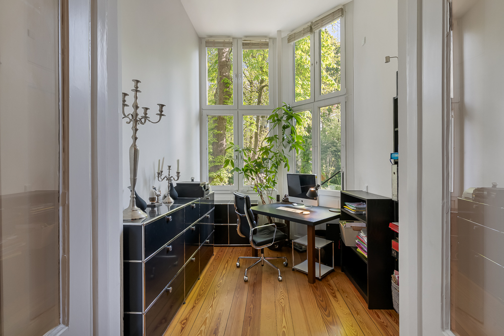 Helles Arbeitszimmer mit Schreibtisch, Computer, Regalen und großen Fenstern mit Blick ins Grüne.